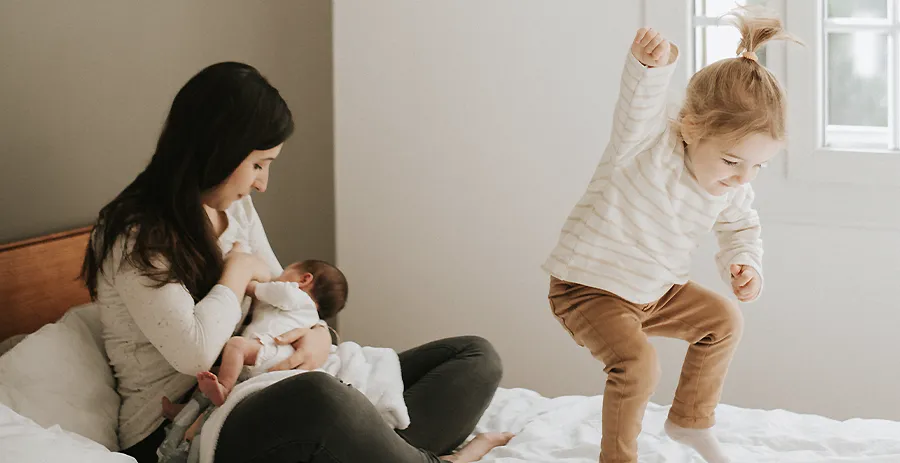 Portrait de famille spontané et en lumière naturelle : une mère allaite son bébé sur le lit tandis que son autre enfant saute à côté.