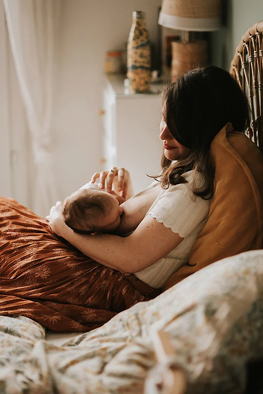 Portrait lifestyle d’une mère qui allaite son bébé dans son lit, en le regardant tendrement avec un sourire complice.