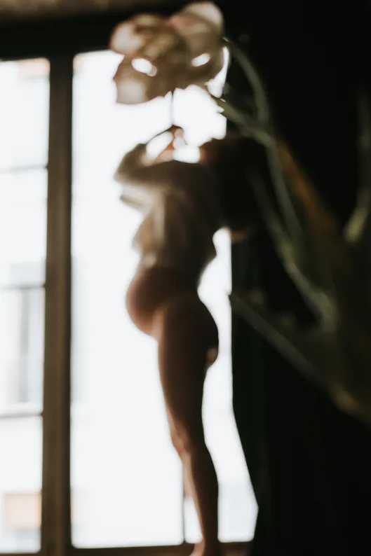 Portrait artistique flou d’une femme devant une fenêtre, surmontée d’une fleur au premier plan, qui semble éclore au-dessus de sa tête.