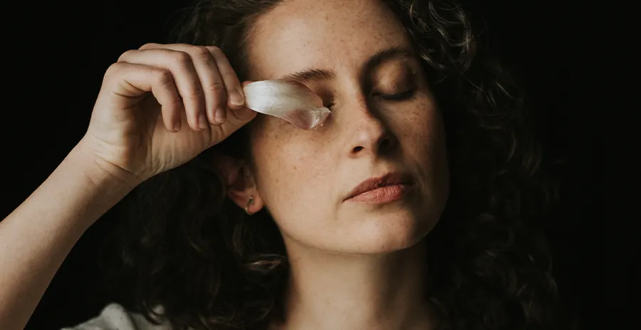 Portrait artistique d’une femme en pull blanc tenant un pétale près de son œil droit, sur un fond noir