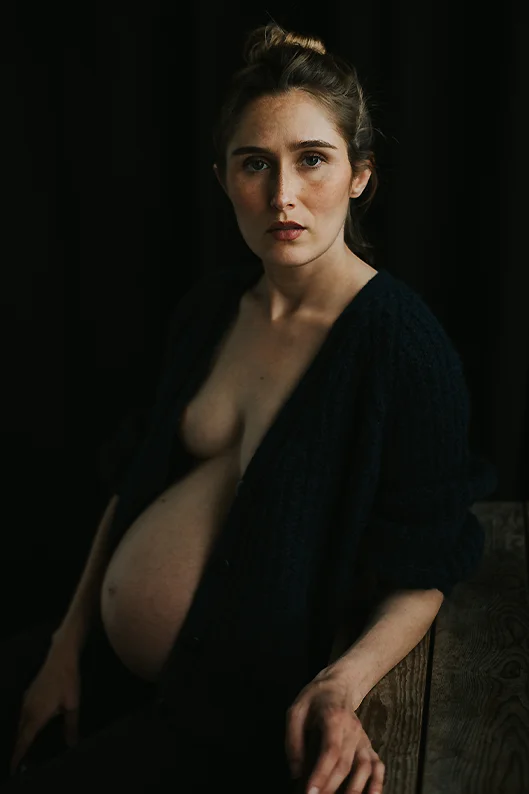 Portrait artistique d’une femme enceinte, vêtue d’un gilet ouvert sur son buste nu, qui pose un regard intense et captivant vers l’objectif.