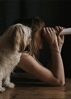 Candice, portraitiste à Paris, allongée sur un parquet ancien, regarde à travers un papier enroulé comme une longue-vue avec sa chienne, Plume.
