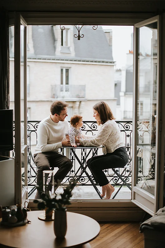 Portrait de famille lifestyle sur un balcon parisien : les parents partagent un moment avec leur bébé, devant les toits de Paris.