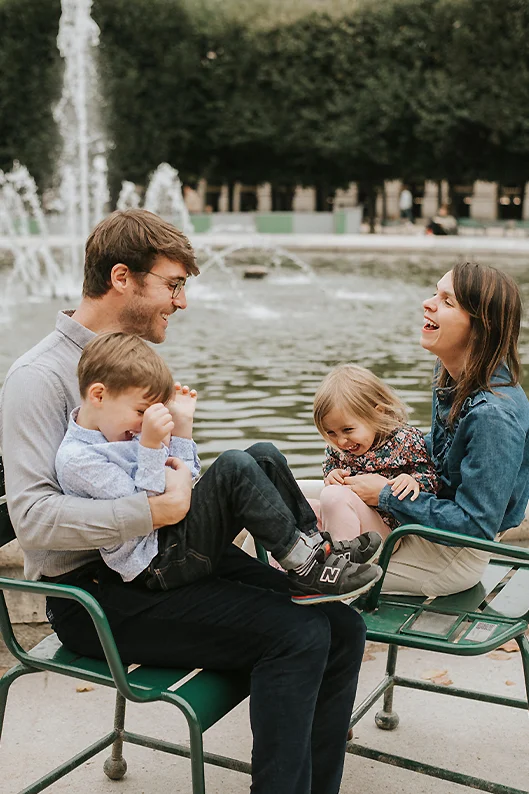 Portrait de famille aux Tuileries : les parents rient avec leurs deux enfants, assis sur leurs genoux, près d’une fontaine.