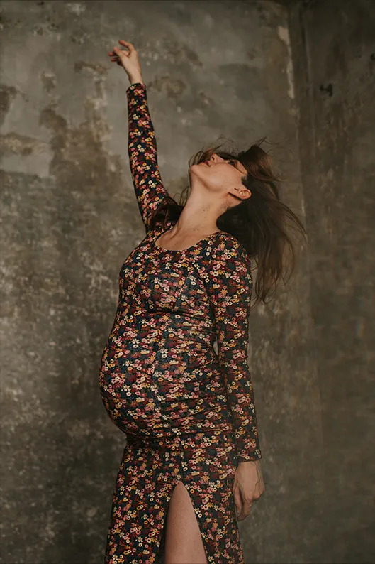 Femme enceinte en robe rose fleurie, un bras levé et les yeux clos, savourant un instant de calme dans une pose artistique.