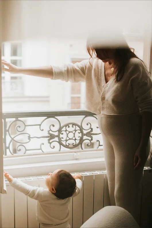 Portrait lifestyle d’une maman et de son enfant près d’une fenêtre, dans une lumière naturelle douce aux tons chauds et enveloppants.
