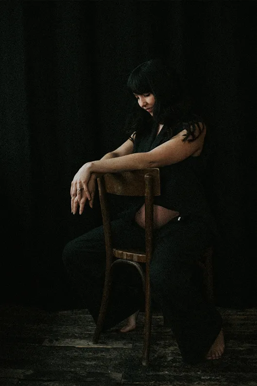 Femme enceinte assise à califourchon sur un tabouret, souriante et pleine d’assurance, dans un portrait de studio à la fois élégant et intime.