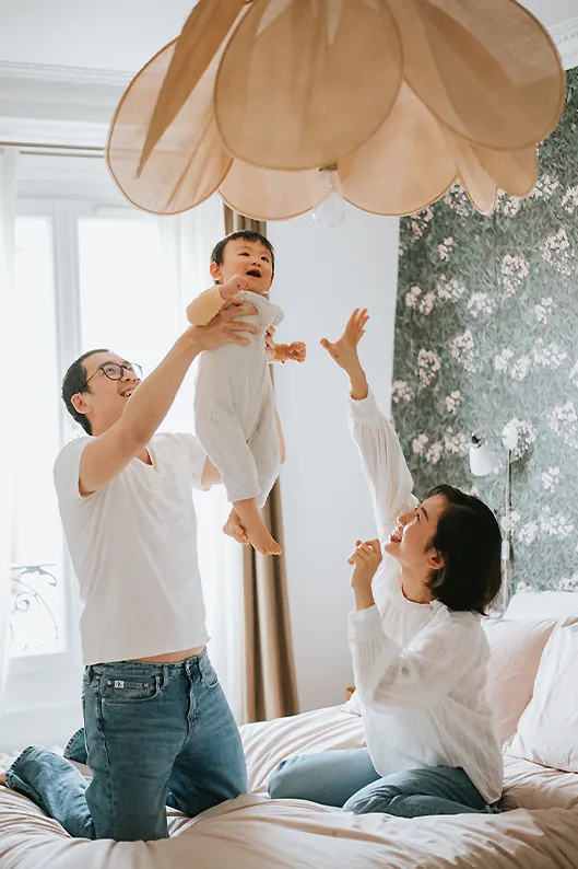 Séance photo de famille à domicile : les parents jouent avec bébé sur le lit en riant.
