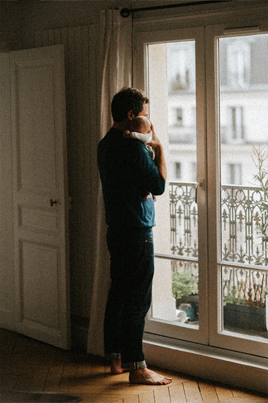Lors d’une séance photo de nouveau-né en famille à domicile, un papa tient son bébé contre lui et regarde au loin par la fenêtre.